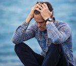 A man in a plaid shirt sits by the water looking distressed, symbolizing stress.